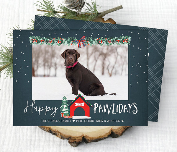 dog themed holiday card with single photo against a blue background. traditional red and green christmas garland along the top and a dog house and christmas tree at the bottom. happy pawlidays wording